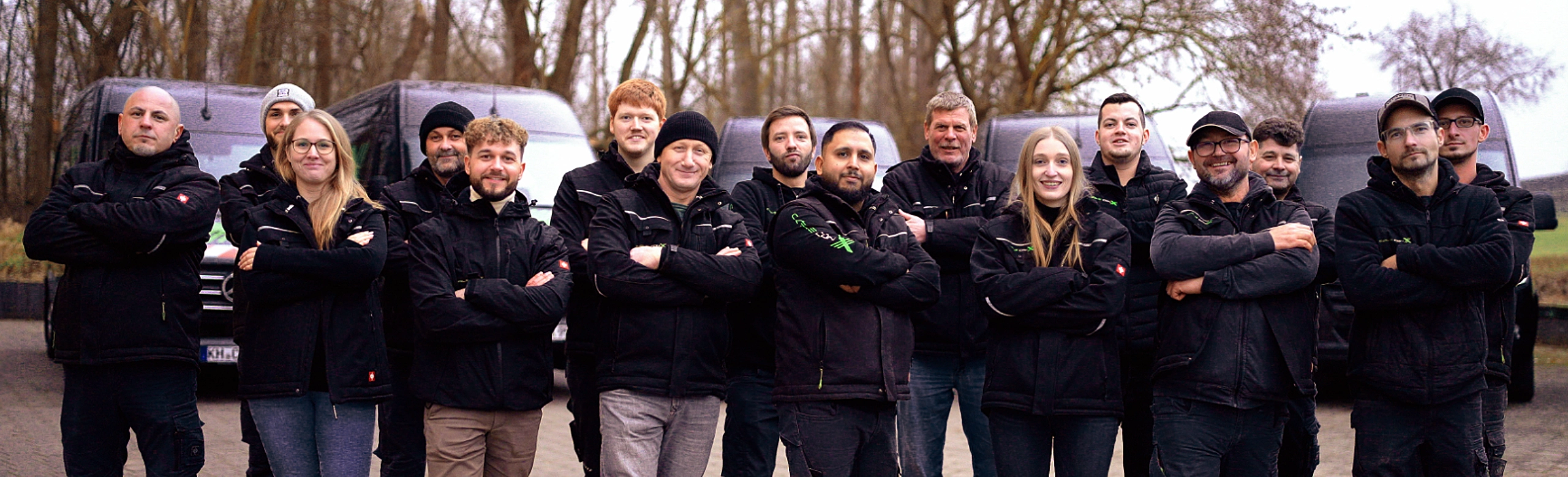 Gruppenbild: 16 Cable-conX-Mitarbeiter in warmer, schwarzer Firmenkleidung stehen vor vier Fahrzeugen der Firmenflotte.